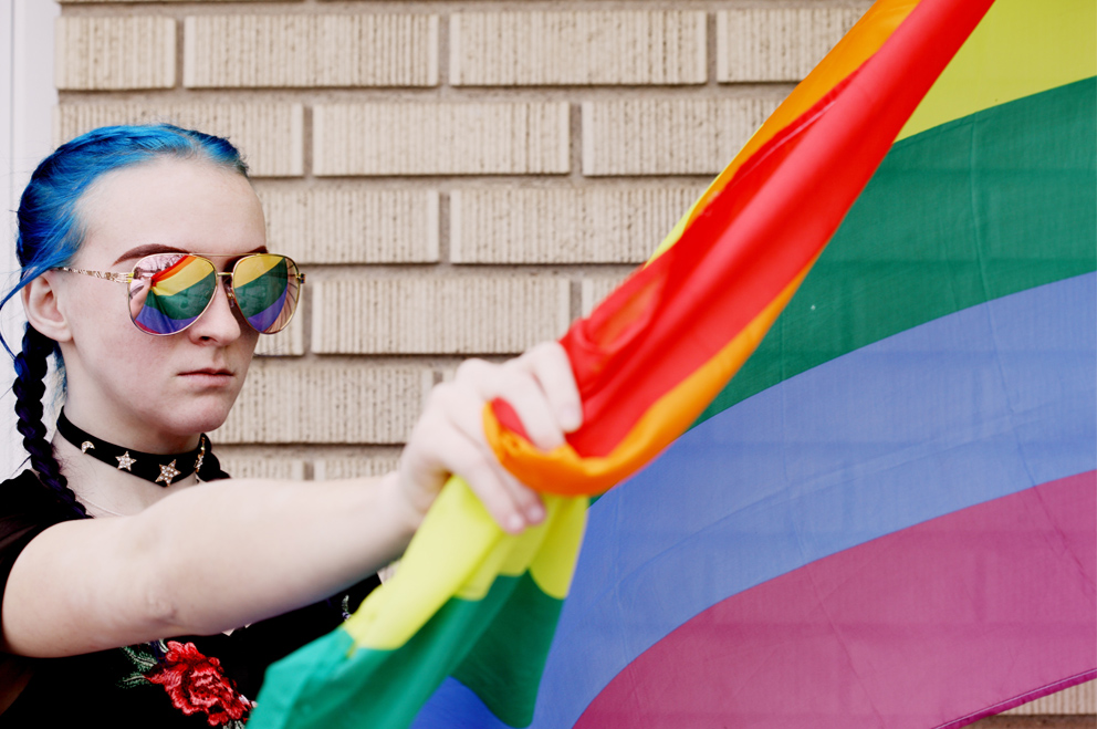 Eine junge Person mit blauen Haaren hält eine Regenbogenflagge, die sich in ihrer Sonnenbrille spiegelt.