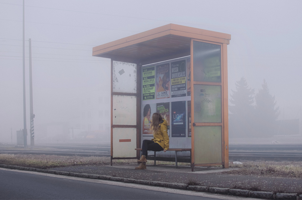 Eine Junge Person sitzt an einer Bushaltestelle im Nirgendwo.