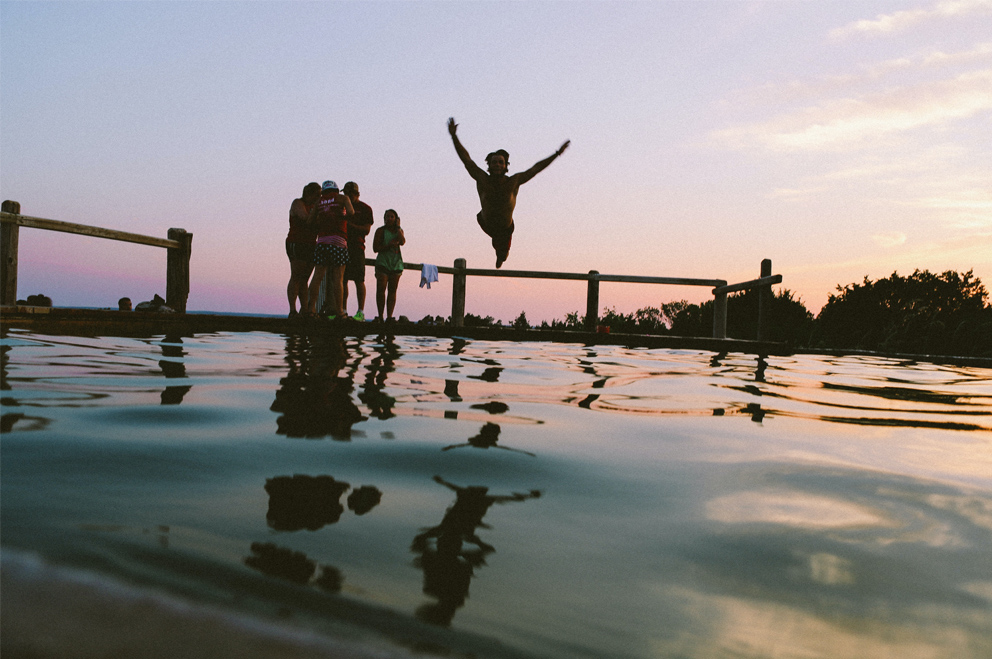 Eine Gruppe junger Menschen an einem See. Eine Person springt mit ausgebreiteten Armen ins Wasser.