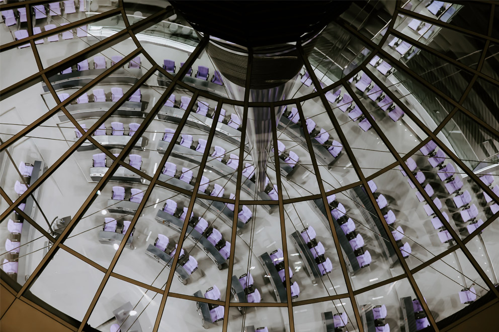 Blick in den menschenleeren Plenarsaal des Deutschen Bundestages durch die Glaskuppel. Im Bild die blauen Stühle und der graue Flur sowie architektonische Merkmale der Kuppel aus Stahl und Glas.