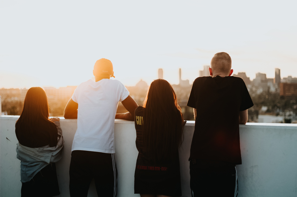 Vier junge Menschen mit dem Rücken zur Kamera lehnen an einer brusthohen weißen Mauer und schauen in die Ferne auf den Sonnenuntergang. Im Hintergrund eine Skyline mit einigen Wolkenkratzern.