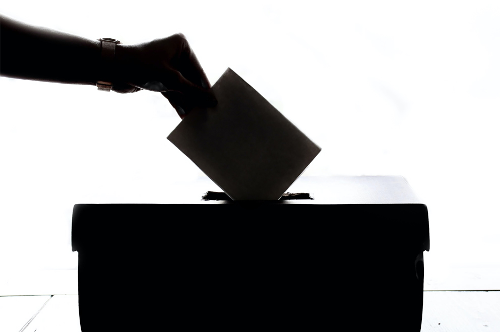 Umrisse einer Hand, die einen Wahlzettel in eine Wahlurne wirft.