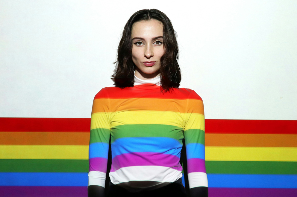 Junge Frau blickt selbstbewusst in die Kamera. Sie trägt einen Pullover in Regenbogenfarben. Auch im Hintergrund ist die Regenbogenfahne im unteren Drittel abgebildet, der restliche Hintergrund ist weiß. 
