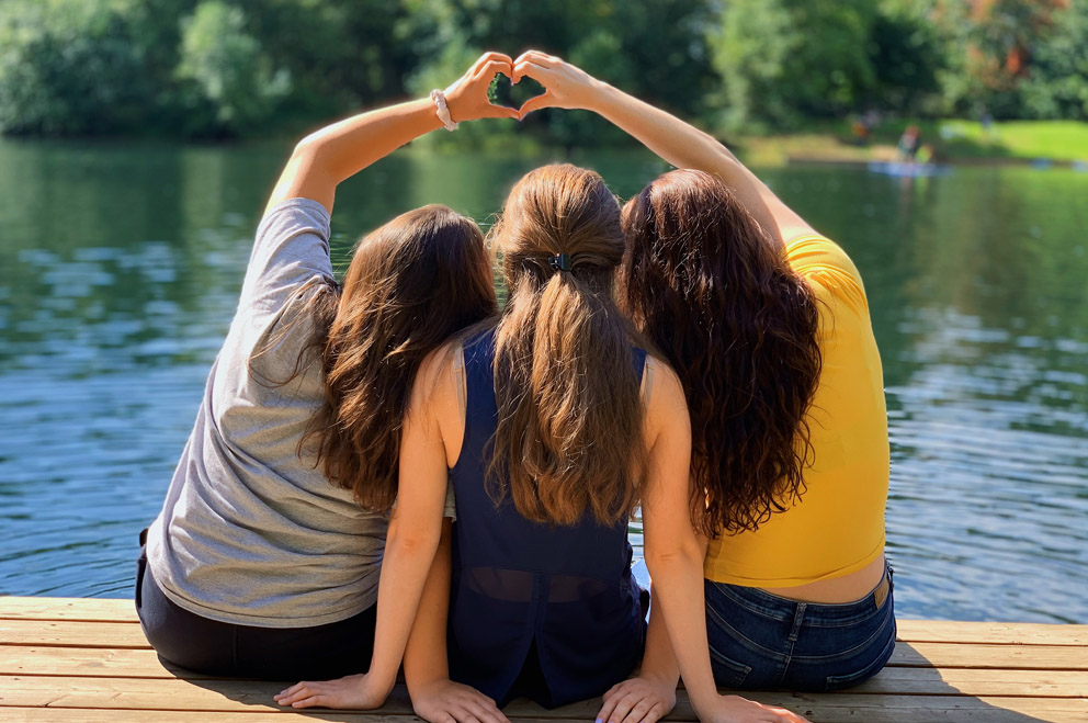 Drei junge Personen mit langen Haaren sitzen auf einem Steg am Wasser. Die beiden äußeren bilden mit ihren Händen ein Herz über der Person in der Mitte.