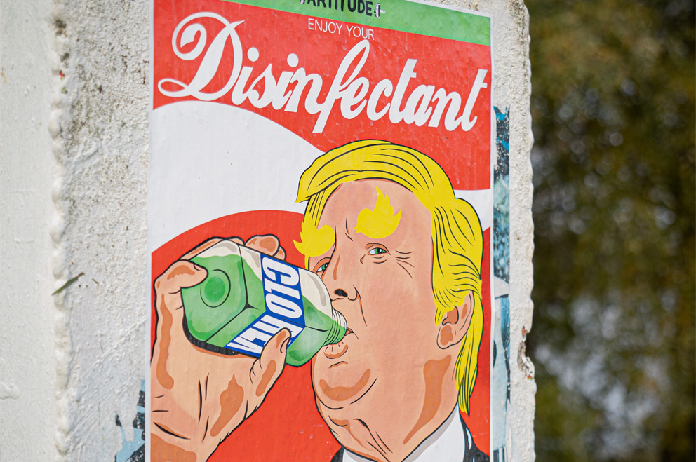 Plakat an einer Wand mit der Zeichnung von Donald Trump, der grüne Flüssigkeit aus einer durchsichtigen Flasche trinkt. Darüber im Design einer bekannten Cola-Marke der Spruch „Enjoy Your Disinfectant“.