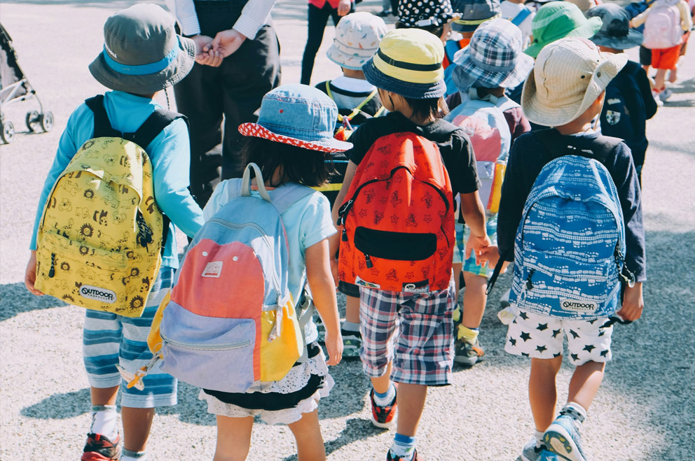 Blick von Hinten auf Gruppe von Kindern im Grundschulalter mit Mützen und Rucksäcken. 