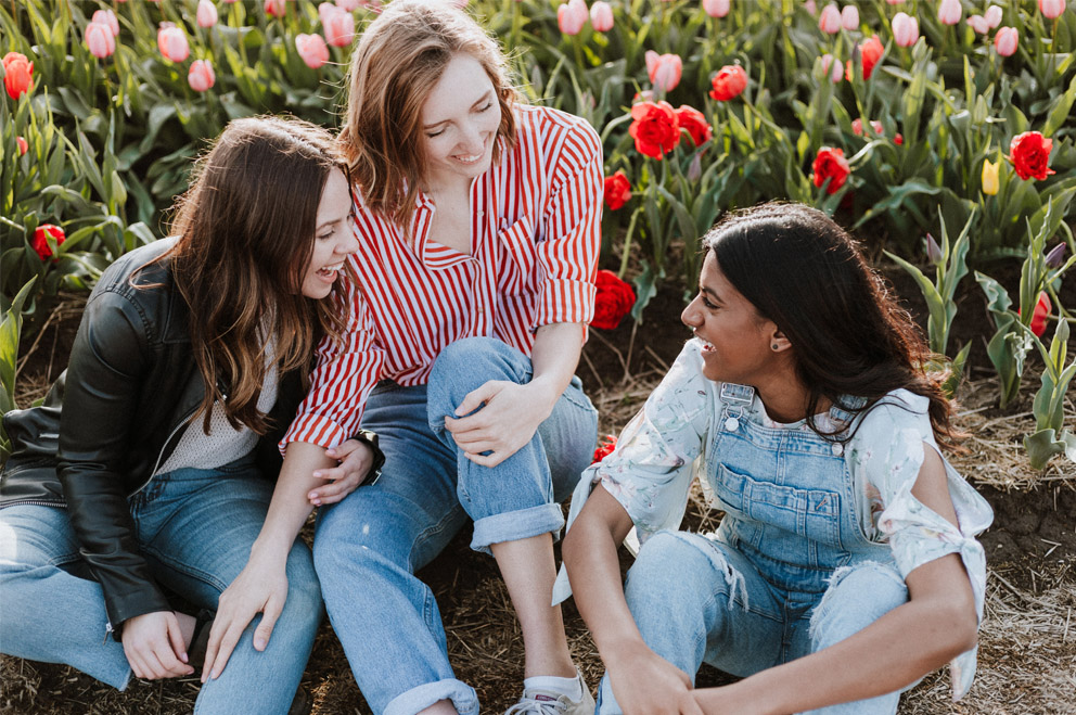 Drei junge Frauen sitzen auf dem Boden und lachen. Im Hitergrund blühende Blumen. Foto: Priscilla Du Preez / Unsplash