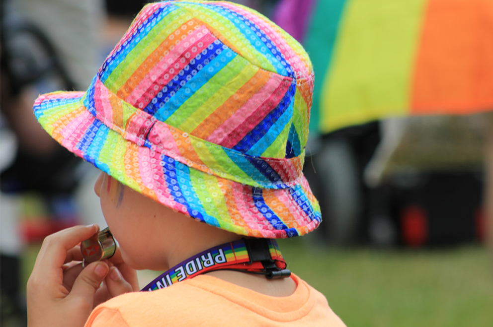 Im Vordergrund ein Kind mit dem Rücken zur Kamera, Anglerhut in Regenbogenfarben und Band in Regenfarben mit Schriftzug „Pride“ um den Hald, das eine Pfeife im Mund hält. Im Hintergrund eine Person mit Regenbogenfahne, deren Unterbeine sichtbar sind.