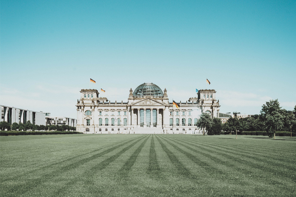 Blick von weitem frontal auf den Reichstag. Davor ein frisch geschnittener Rasen, darüber ein wolkenloser blauer Himmel. 