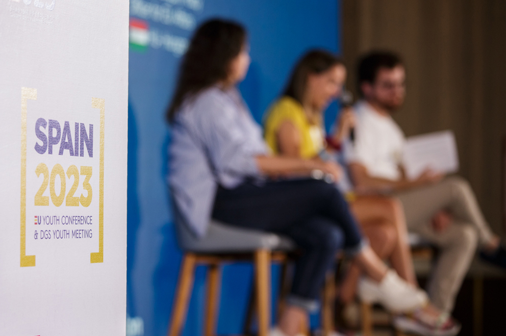 Schild im Vordergrund mit Schriftzug "SPAIN 2023. EU Youth Conference and DGS Youth Meeting". Im Hintergrund unscharf drei diskutierende Menschen auf Hockern sitzend.