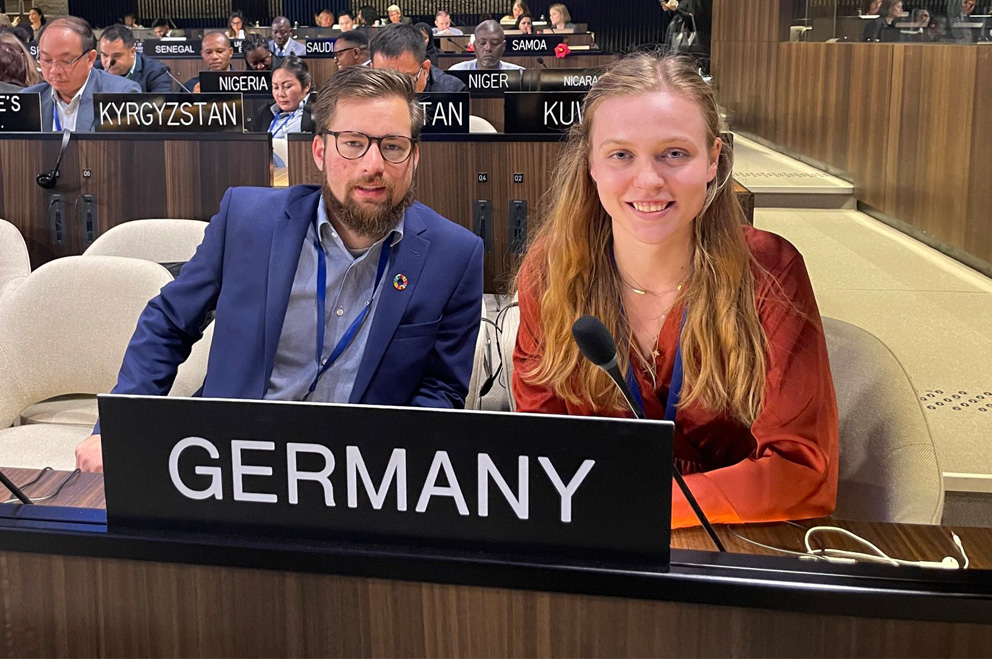 Die beiden Jugenddelegierten für Nachhaltige Entwicklung sitzen im Verhandlungssaal hinter einem Schild mit der Aufschrift "Germany". 