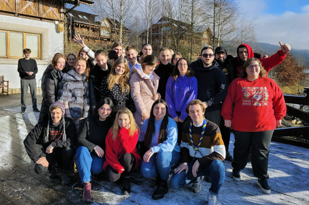 About 25 young people pose for a group picture on a terrace during the networking meeting of the National Youth Council of Ukraine. In the background, you can see half-timbered buildings on the left, barren trees in the middle and some clouds in the sky. To the right, hills and a valley can be seen in the background. 