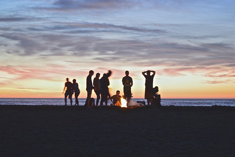 Jugendliche stehen um ein Lagerfeuer am Strand.