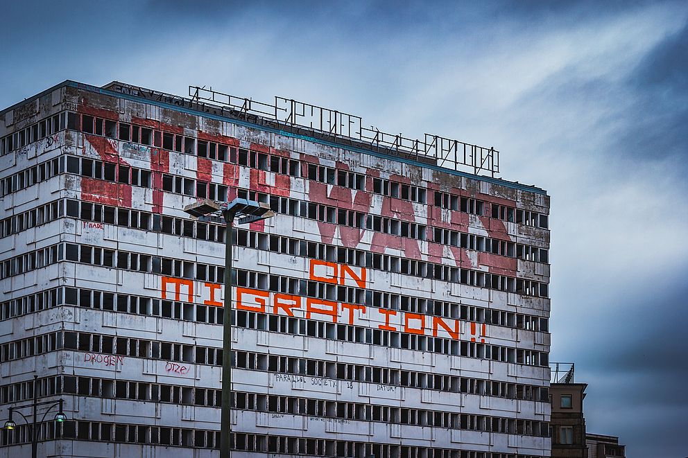 Ungenutztes Hochhaus mit ausgeschlagenen Fenstern am Berliner Alexanderplatz mit einem großen Graffiti in roter Schrift „Stop Wars in Migration“ (Stoppt die Kriege gegen Migration). 